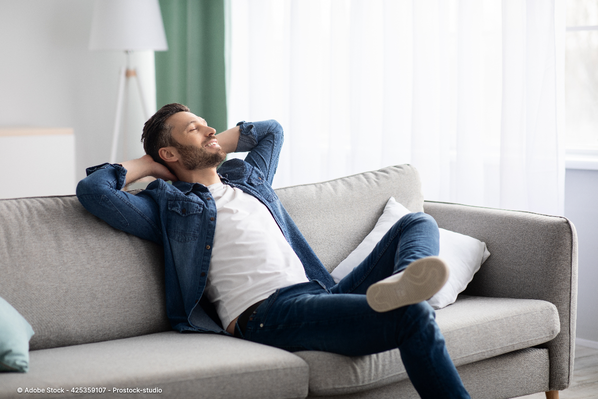 Ein entspannter Mann liegt lächelnd auf einem grauen Sofa und veranschaulicht Zuhause wohlfühlen Tipps für gemütliche Stunden.